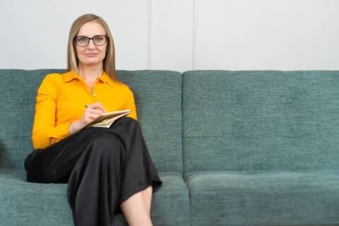 Beautiful young psychotherapist takes notes, looks into camera and smiles while sitting on couch in office. Portrait beautiful female psychologist with glasses, posing with clipboard in room