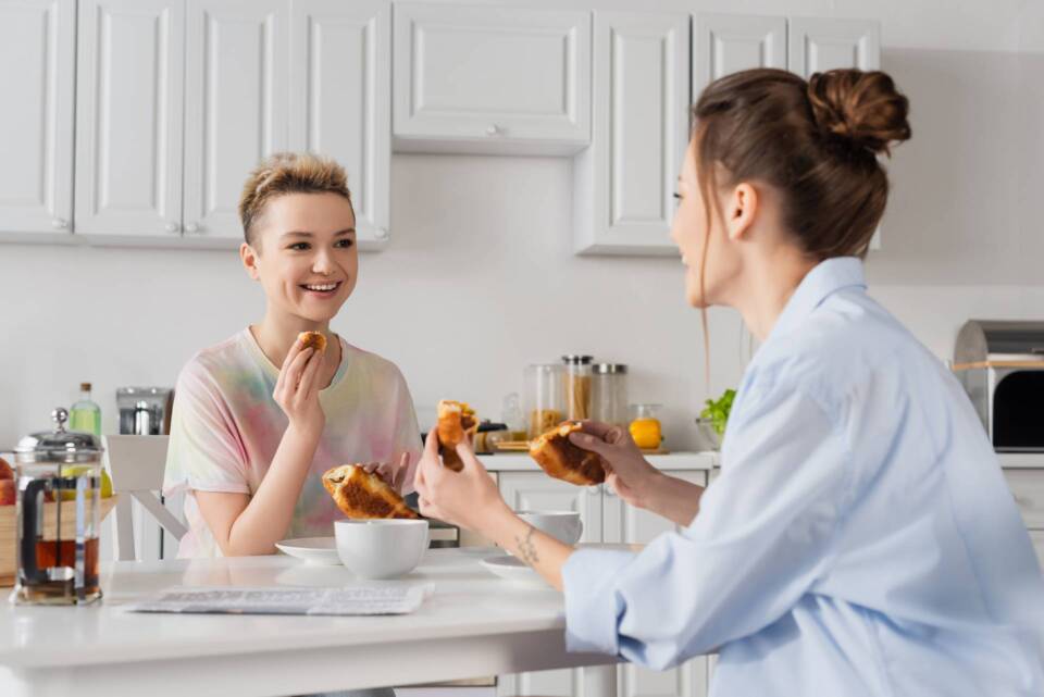 Joyful pansexual couple eating delicious croissants for breakfast.